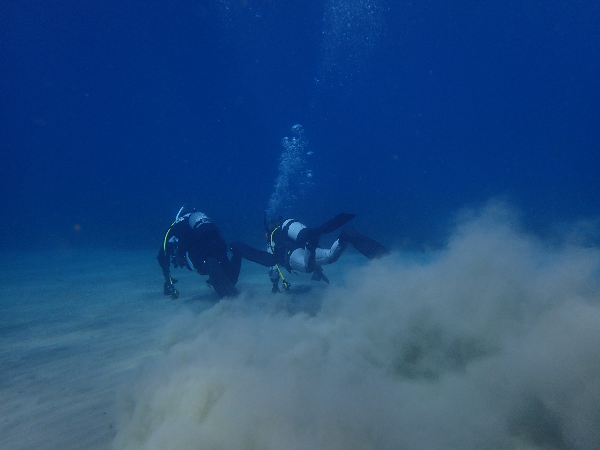 青い海で、水底付近を泳ぐ２人のダイバーが、水底の砂をモクモクと巻き上げながら去ってゆく後ろ姿
